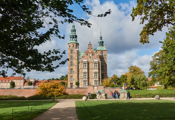 The image features Rosenborg Castle, a notable example of Renaissance architecture located in Copenhagen. Captured during the late morning in autumn, the photograph showcases the castle with its distinctive towers and intricate brickwork, set against a partly cloudy sky. The castle is surrounded by lush gardens with green grass and trees showing seasonal autumn colors. Visitors are visible in the foreground, adding scale to the scene, while the castle serves as the prominent landmark. This is an architecture photograph taken in Copenhagen that highlights both the historical significance and the well-preserved design of Rosenborg Castle in an autumnal setting.